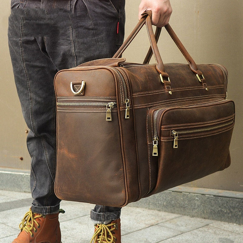 Brown leather suitcase held by a person wearing dark jeans and brown boots.