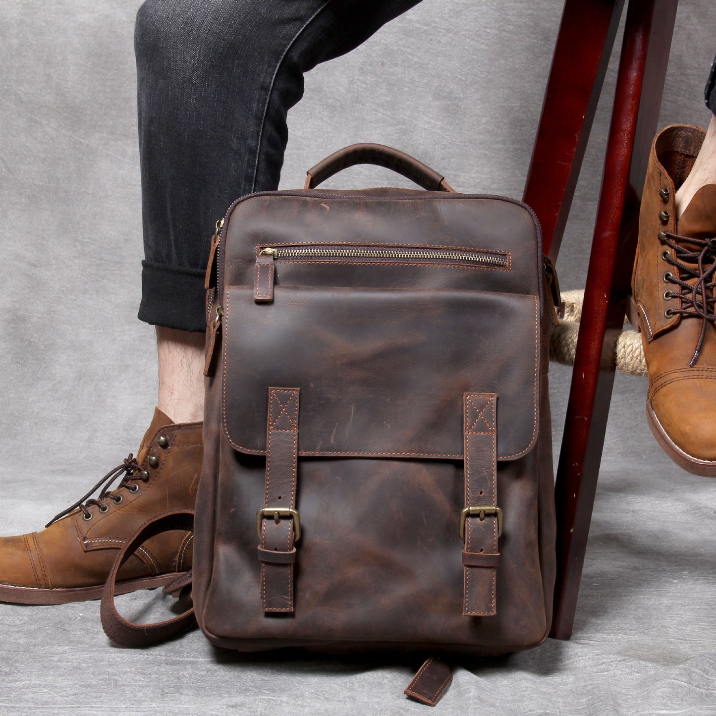 Brown leather backpack on a gray surface with a person wearing brown shoes.