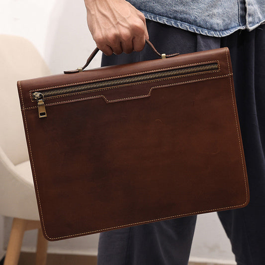 Brown leather briefcase held by a person wearing jeans and a denim jacket.